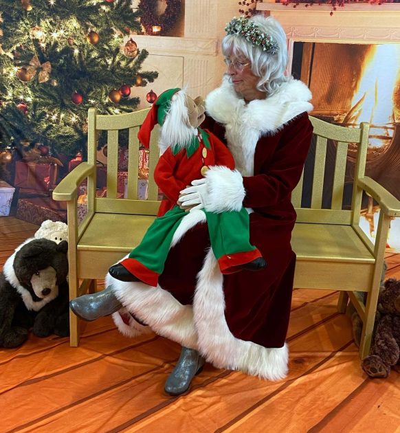 Santa Claus sitting on a bench holding a baby dressed as an elf.