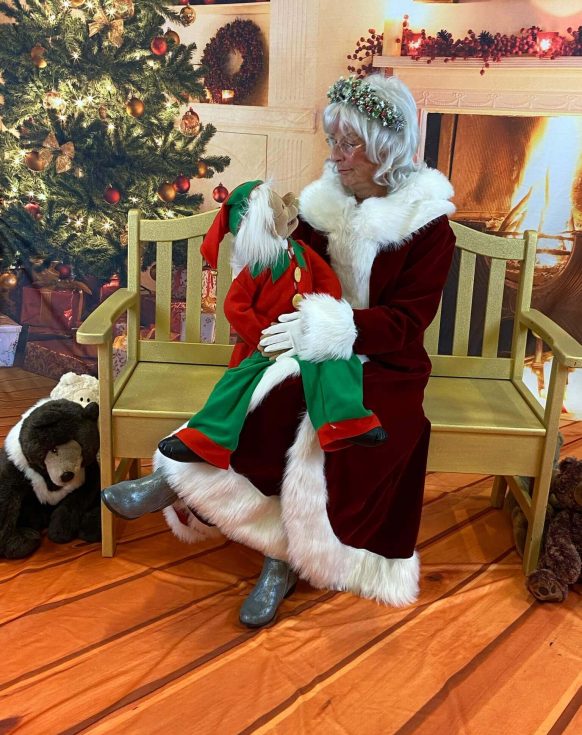Santa Claus sitting on a bench holding a baby dressed as an elf.