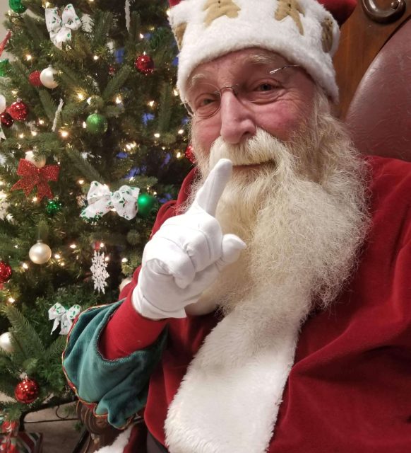 Santa Claus giving a shushing gesture in front of a decorated Christmas tree.
