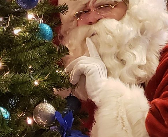 Santa Claus with finger to lips beside Christmas tree.