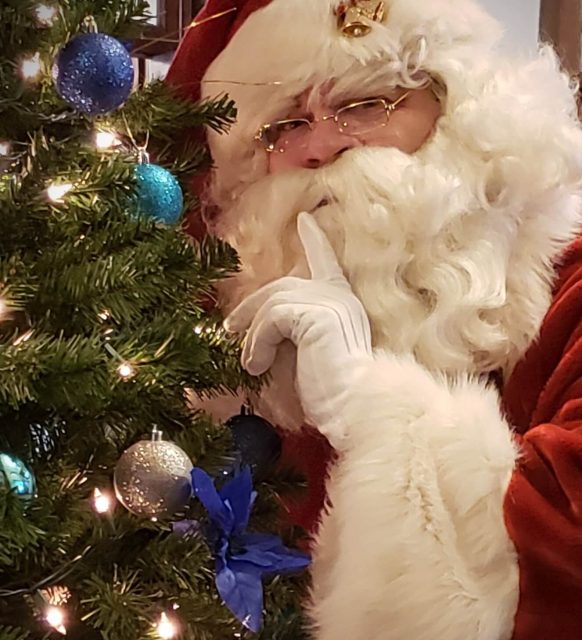Santa Claus with finger to lips beside Christmas tree.